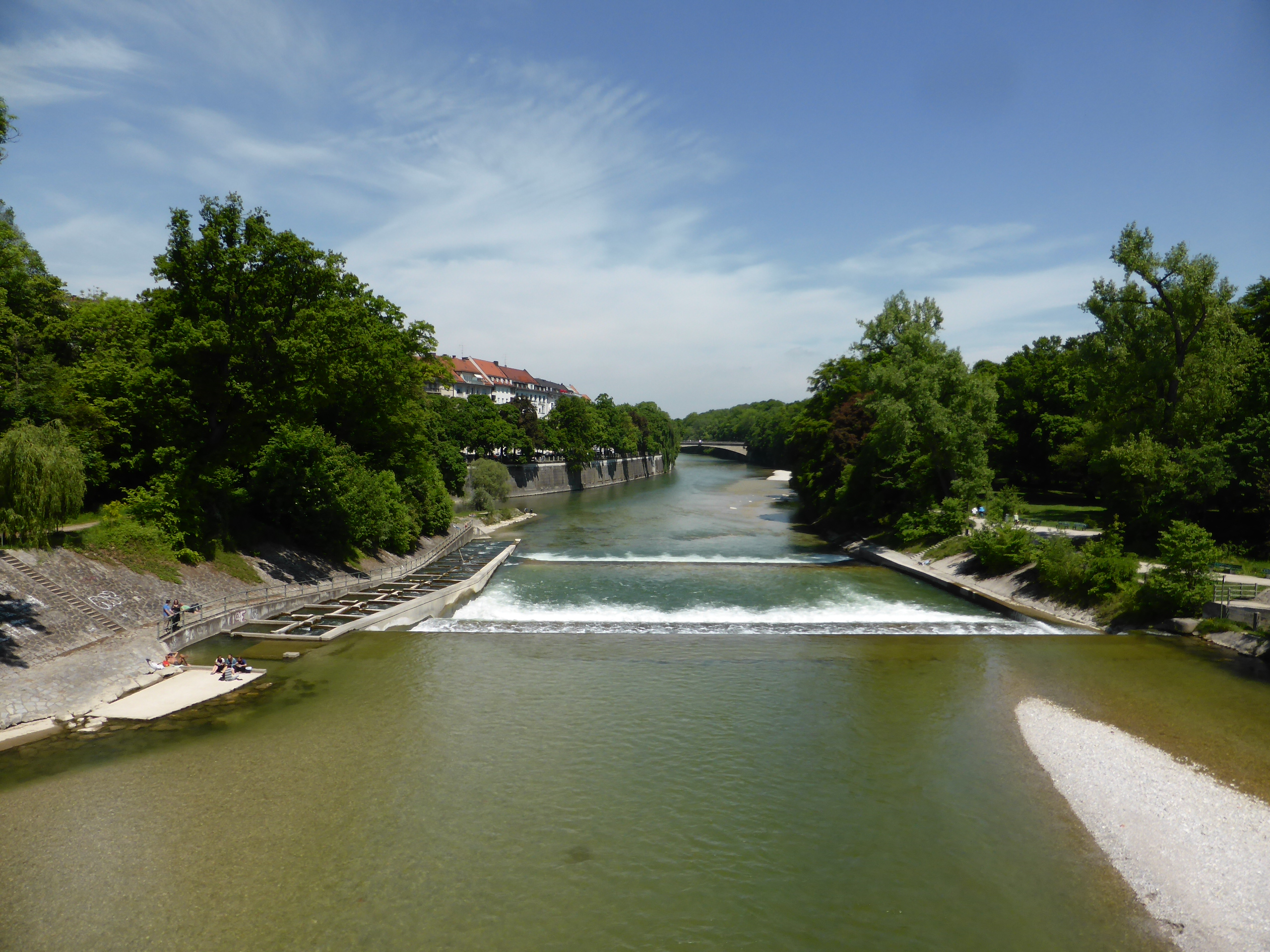 El río Isar atravesando Múnich entre árboles, taludes de piedra y un pequeño azud.