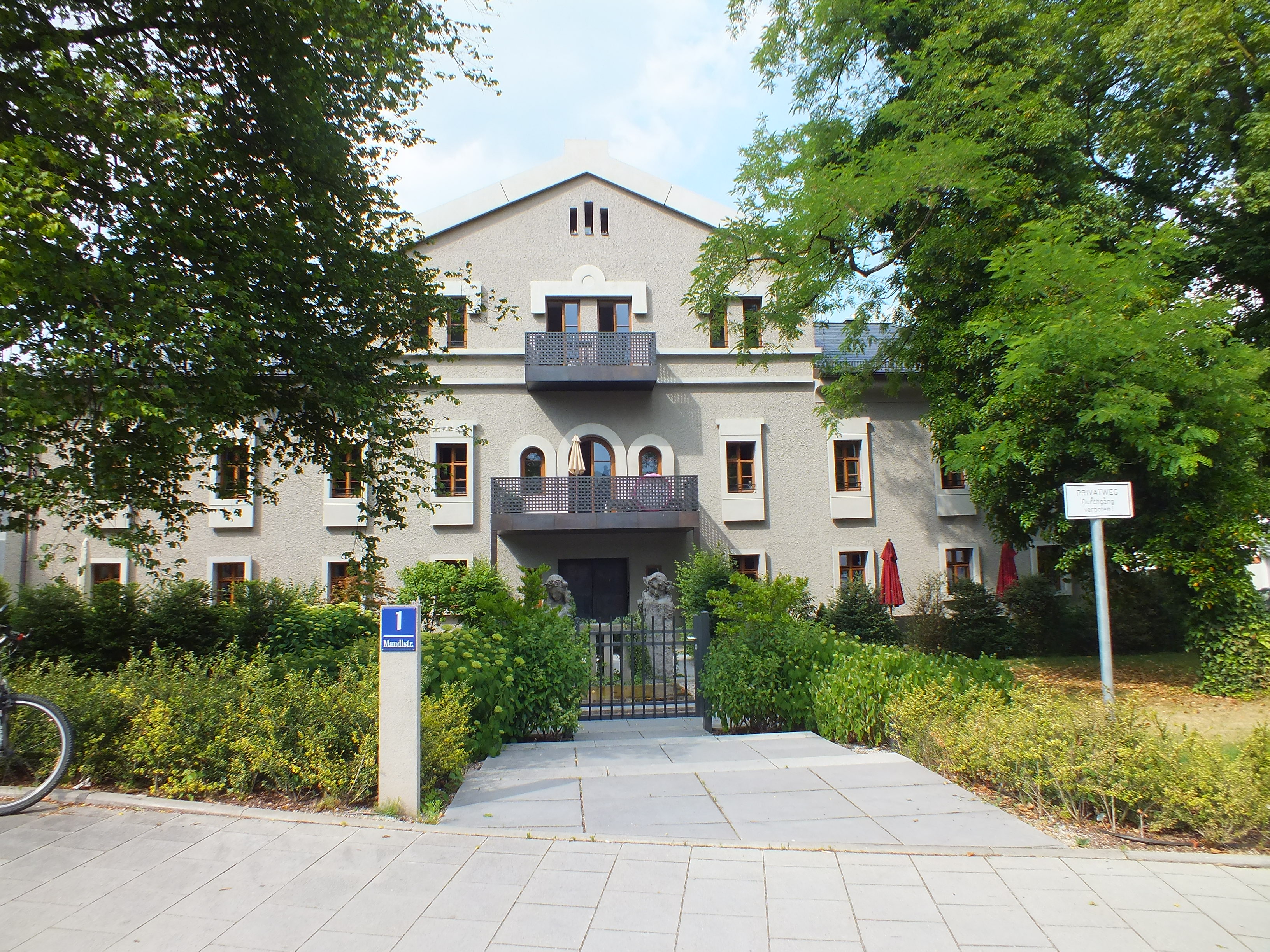 Casa de fachada gris con jardín y placa azul de Mandlstraße en el barrio muniqués de Schwabing.